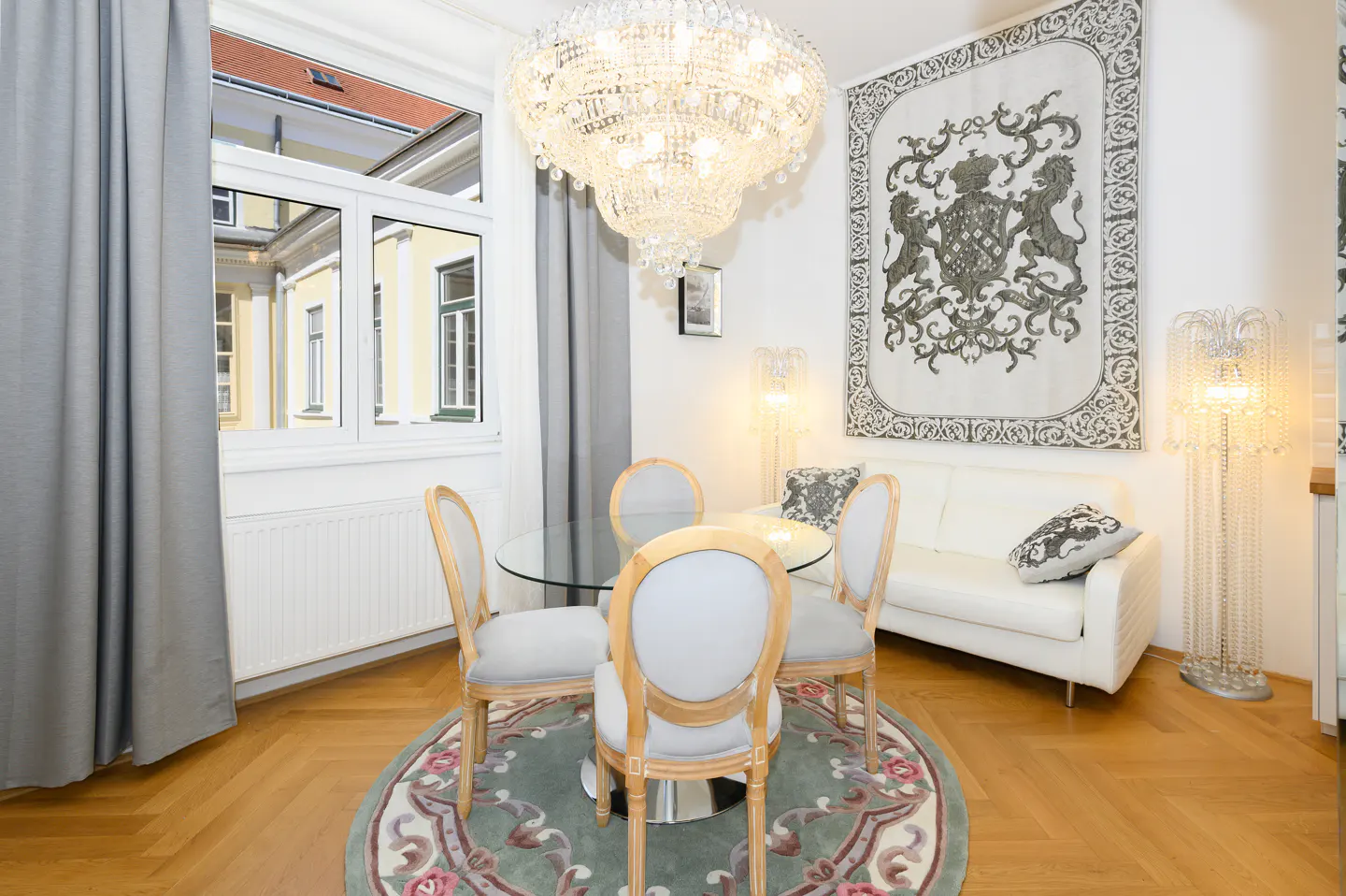 Elegant dining area with a glass table, four chairs, and a crystal chandelier. A white sofa and tapestry are in the background.