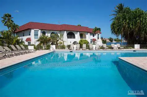 A bright blue swimming pool sits in front of a white building with a red tile roof. Lounge chairs line the pool deck. Palm trees and a clear blue sky are in the background.