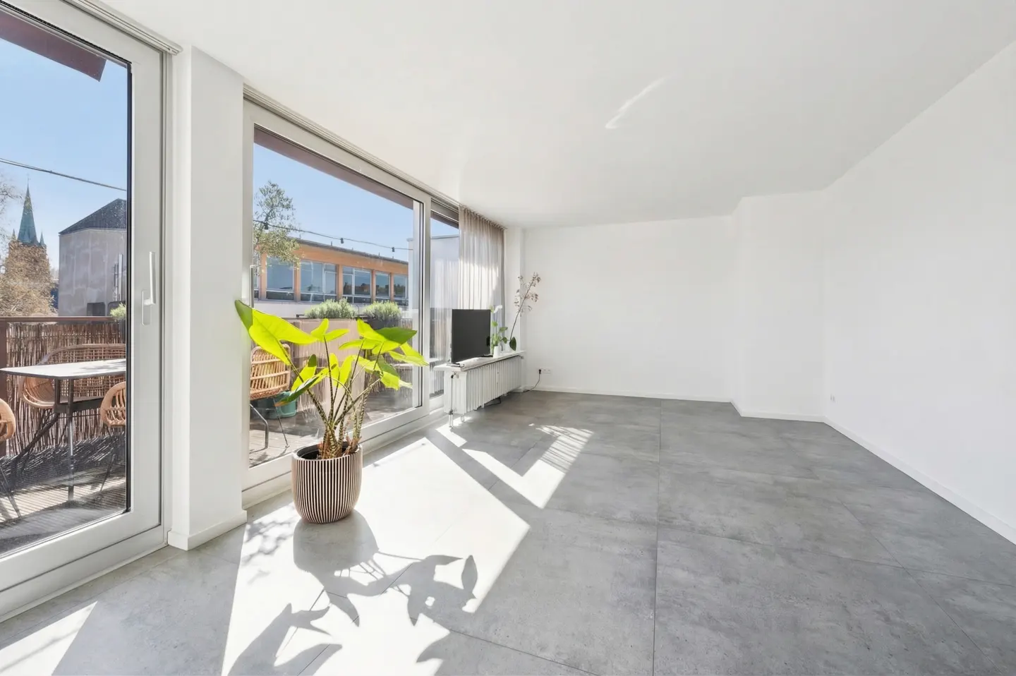 Bright, empty room with gray floors, white walls, and large windows leading to a balcony with outdoor furniture. A potted plant sits near the window.
