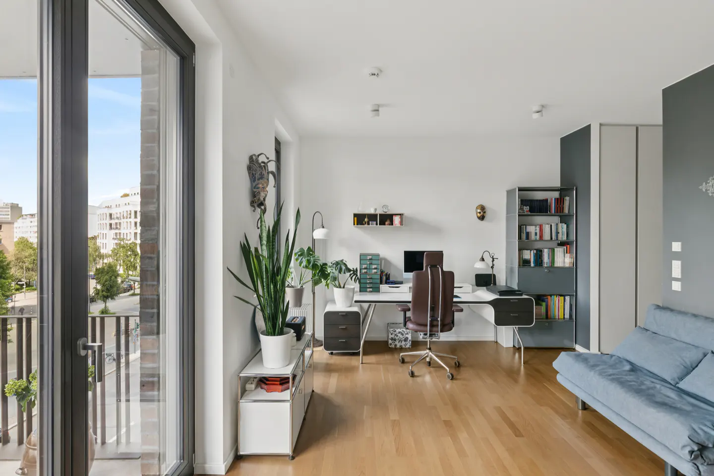 Modern home office with a white desk, brown chair, tall bookshelf, and a blue sofa on hardwood floors near a balcony door.