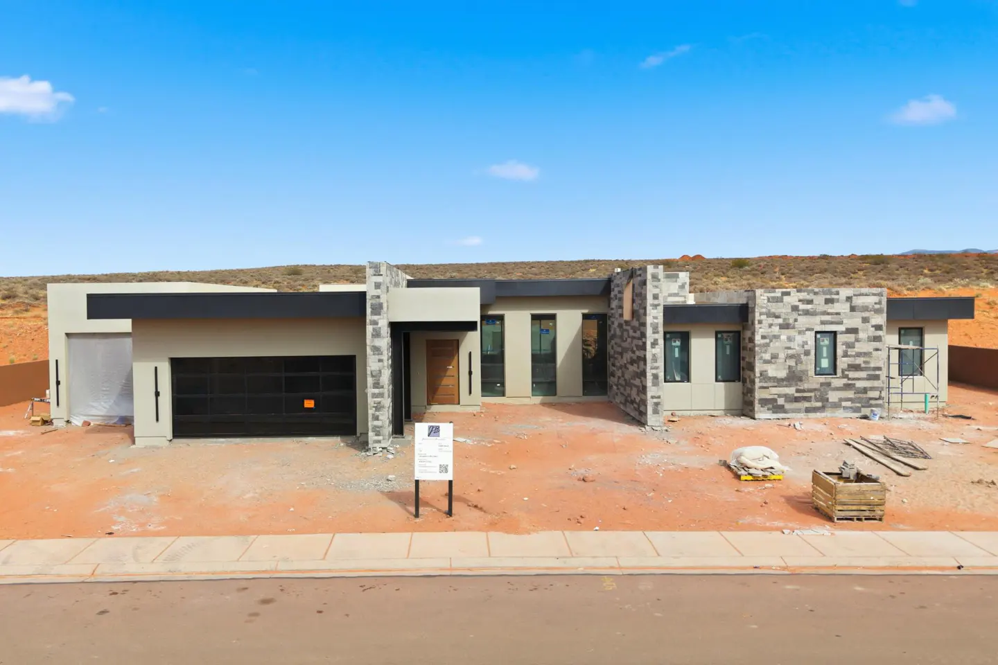 A modern, single-story home with a black garage door and stone accents, set against a blue sky and red dirt landscape.
