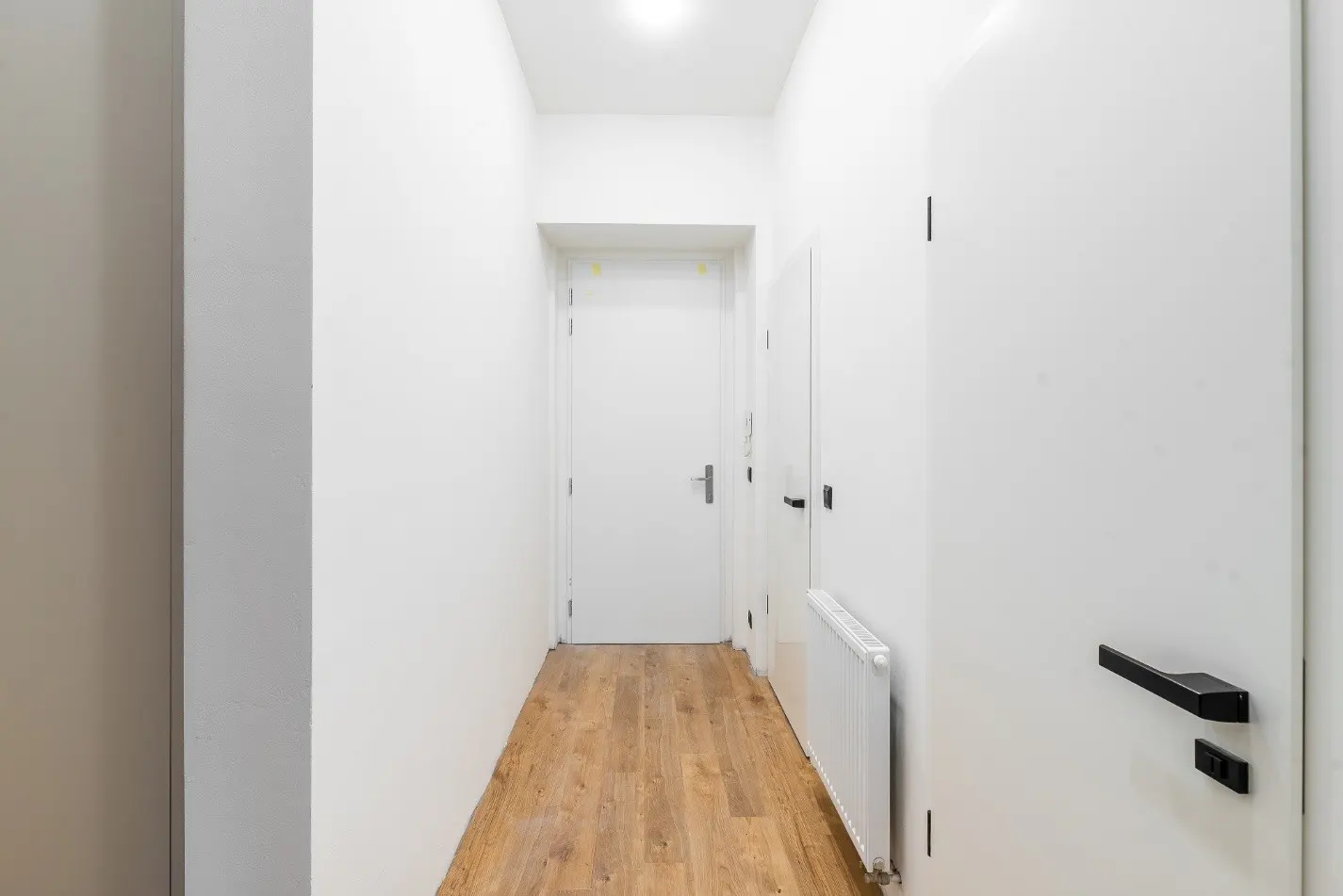 Bright hallway with wood floors, white walls, and white doors with black handles. A radiator is visible along the wall.