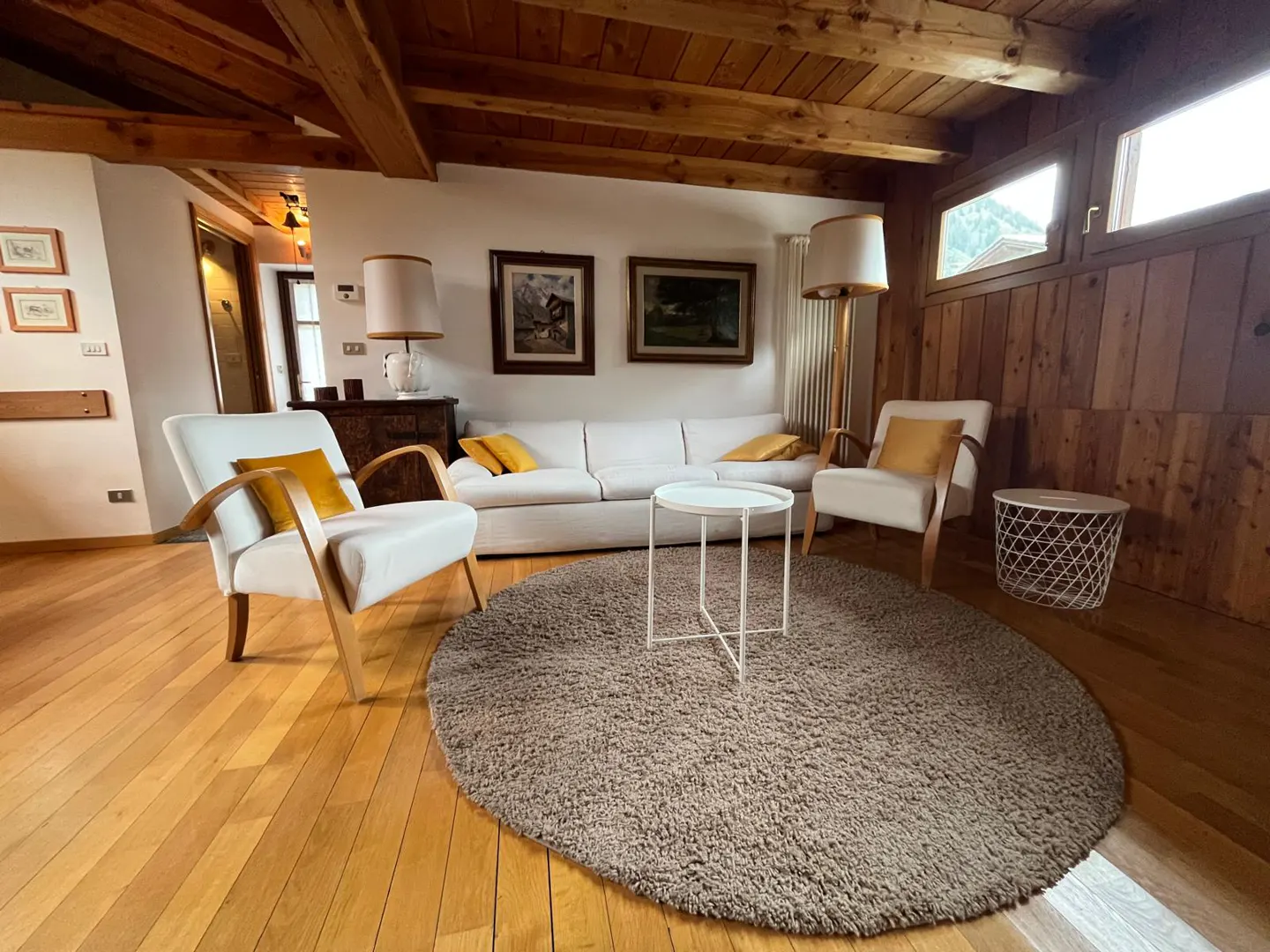 Cozy living room with white sofa and armchairs, round rug, wooden ceiling beams, and warm lighting in a rustic home.