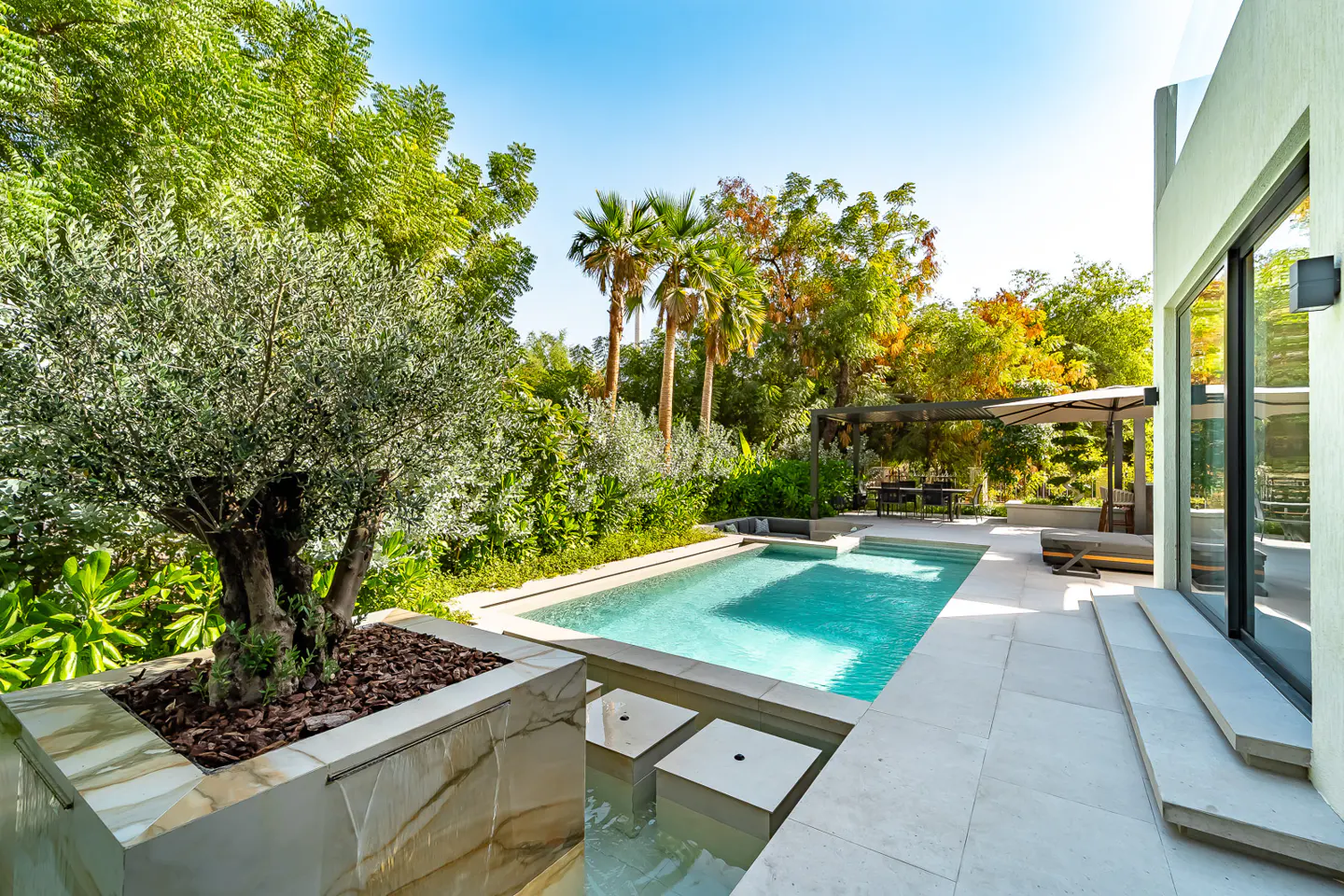 A backyard pool with turquoise water, a stone patio, and lush green trees under a blue sky.