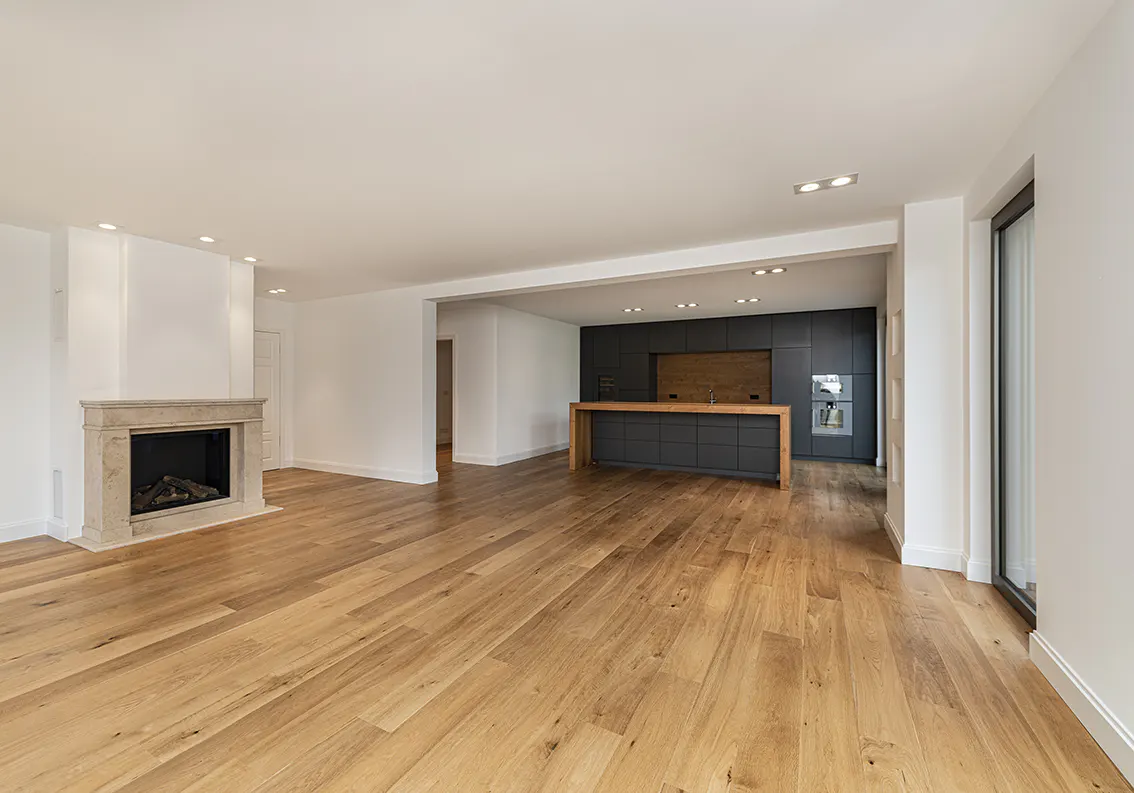 Open-concept living space with light wood floors, white walls, and a stone fireplace. A modern kitchen with dark cabinets is visible in the background.