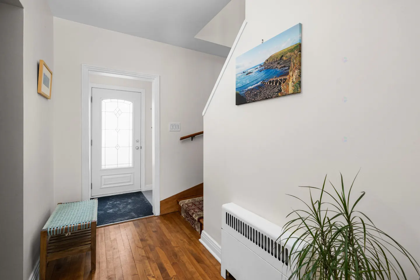 Foyer with hardwood floors, white walls, and a white front door with glass panels. A bench sits near the door, and a seascape painting hangs above a radiator.