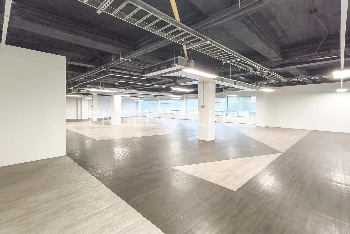 Empty commercial space with white walls, gray flooring, and large windows. Overhead lighting and exposed ceiling fixtures.