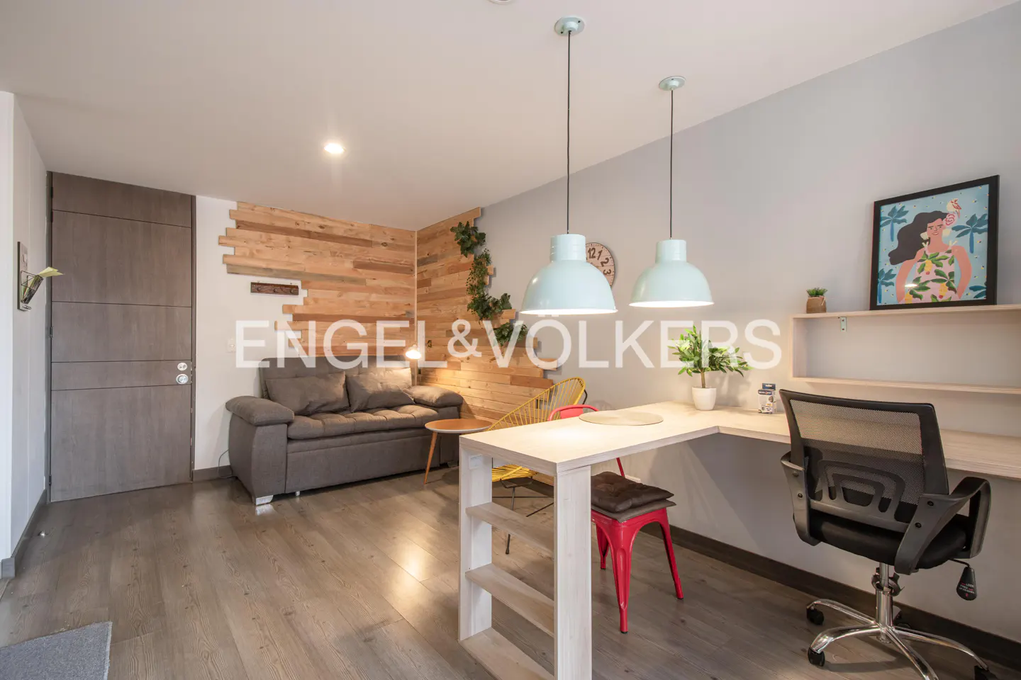 Interior view of a modern apartment with gray sofa, wooden wall, desk, and hanging lights. Engel & Volkers logo is visible.