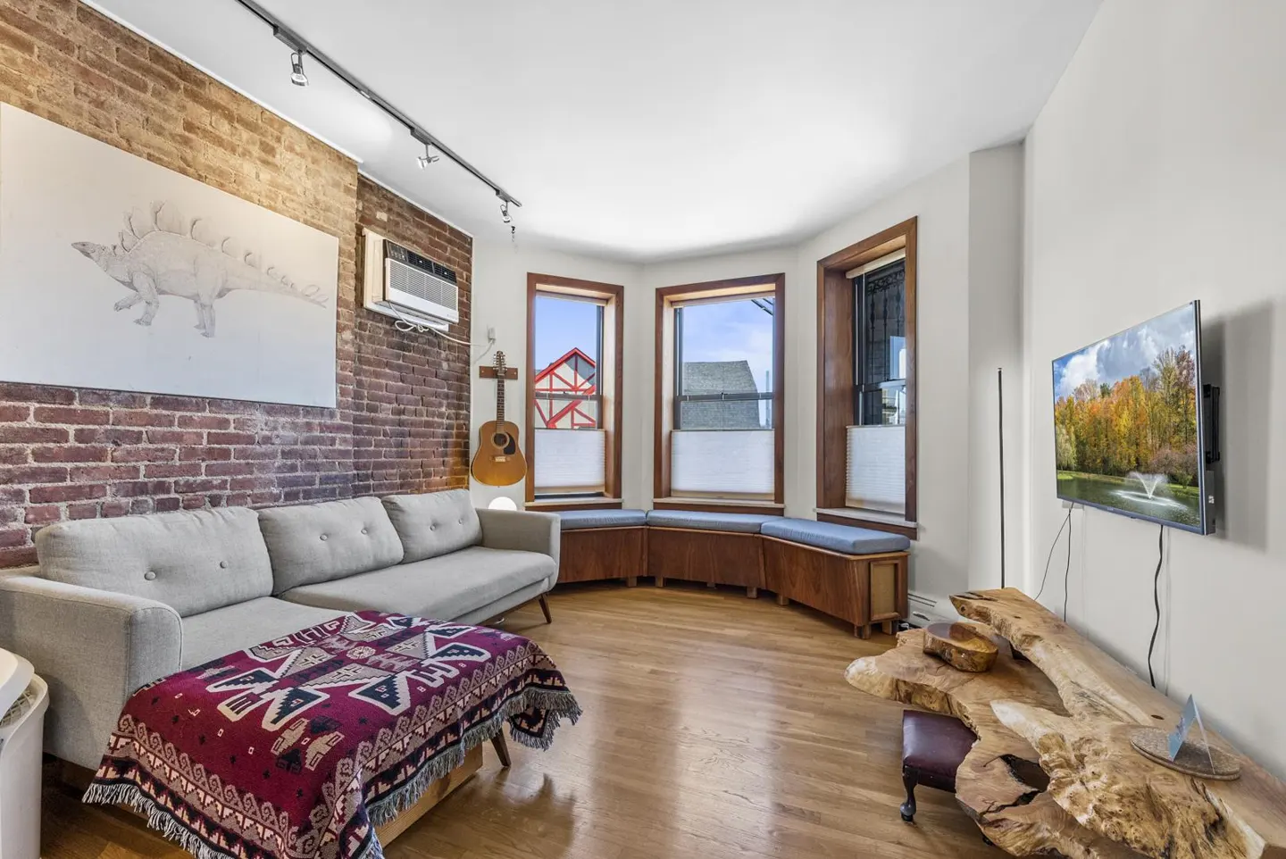 Living room with exposed brick wall, gray sofa with patterned blanket, and bay window with built-in seating. A guitar hangs on the wall.