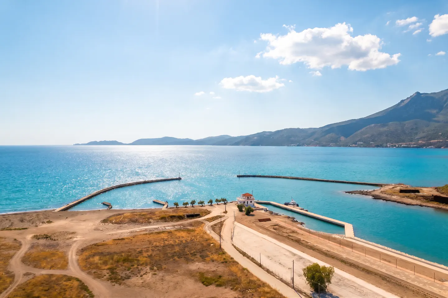 Aerial view of a coastal landscape with turquoise water, sandy beaches, and a small white building near a harbor entrance.