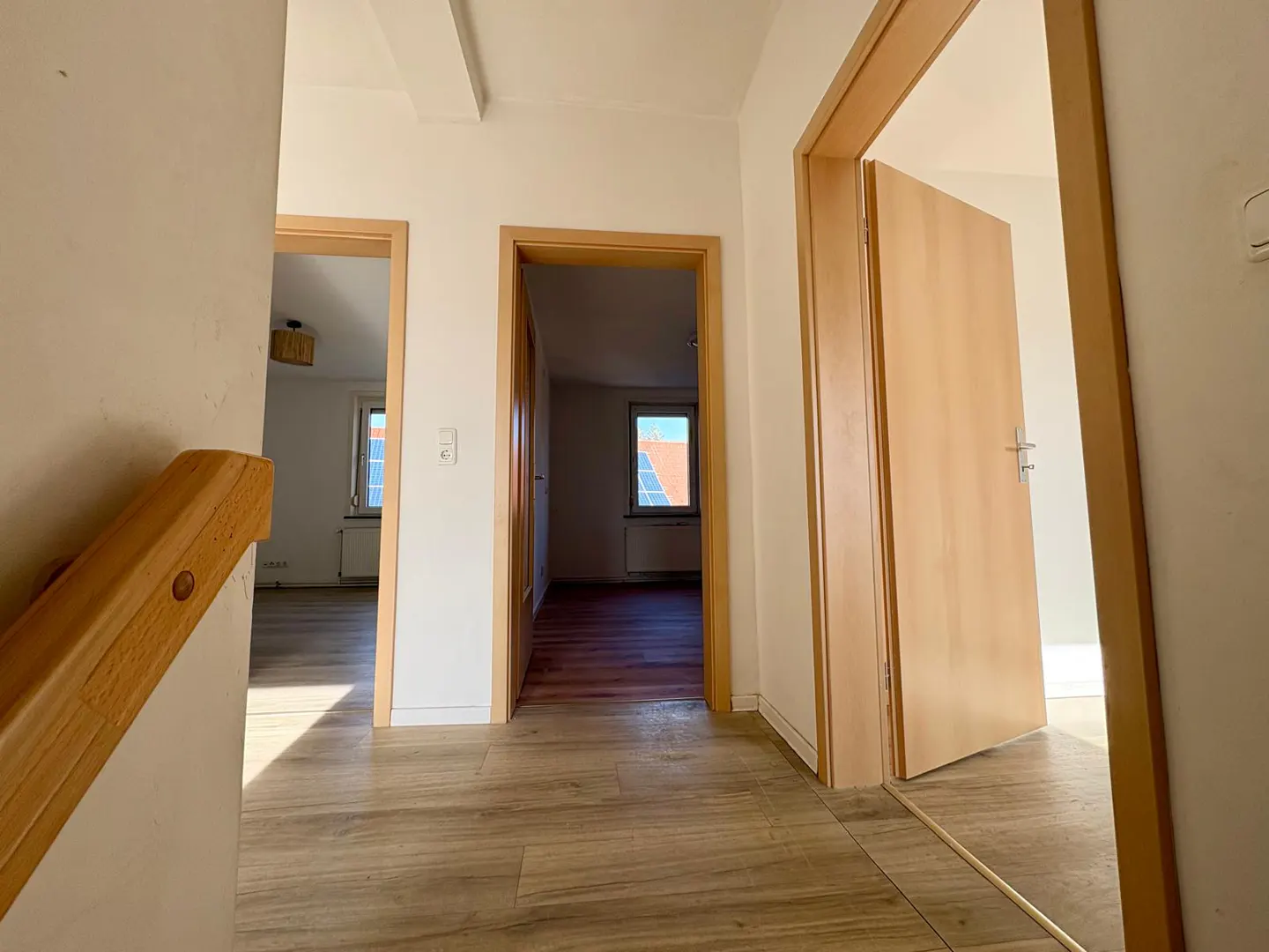 Hallway with light wood floors, white walls, and three open doorways leading to rooms with windows. A wooden handrail is visible.