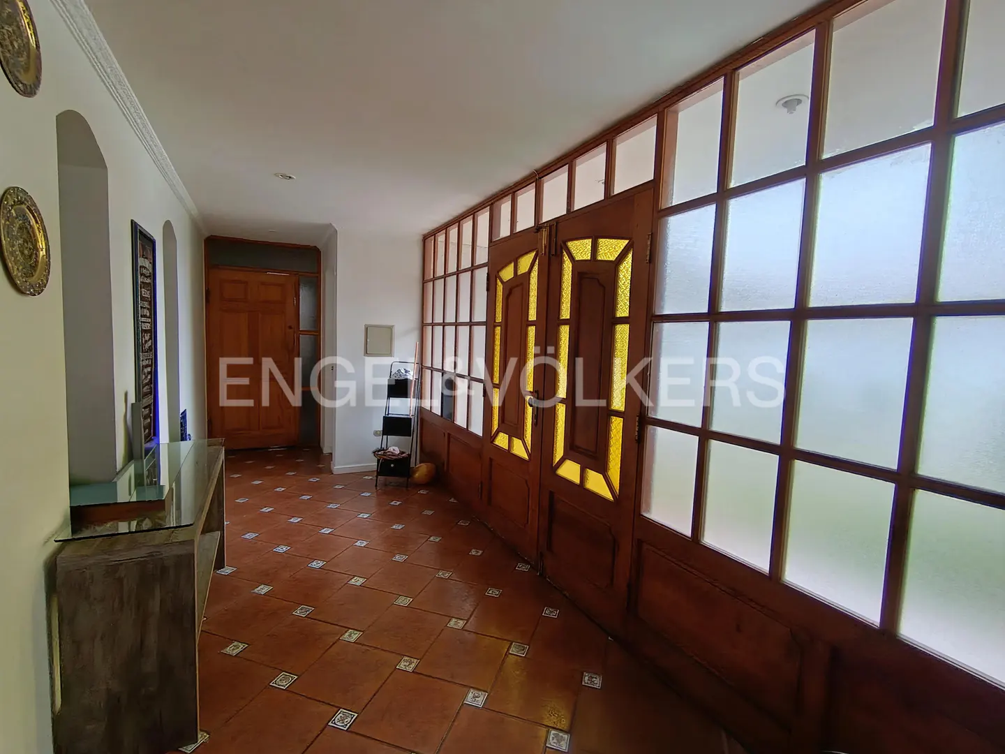 A hallway with terracotta tile floors, a wooden console table, and a large wooden door with yellow glass panels.