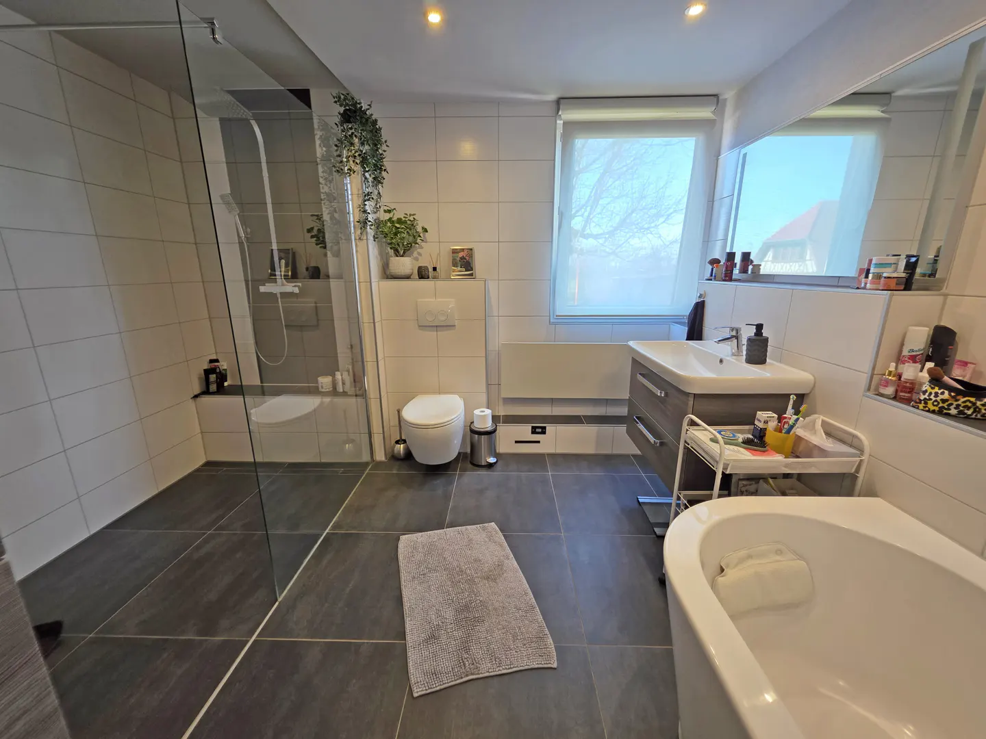 Bright bathroom with gray tile floor, white walls, glass shower, toilet, sink, and white bathtub. A gray rug is on the floor.