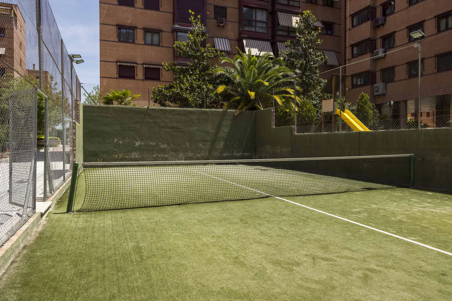 Outdoor padel tennis court with green turf, net, and walls. Apartment buildings and trees are in the background.