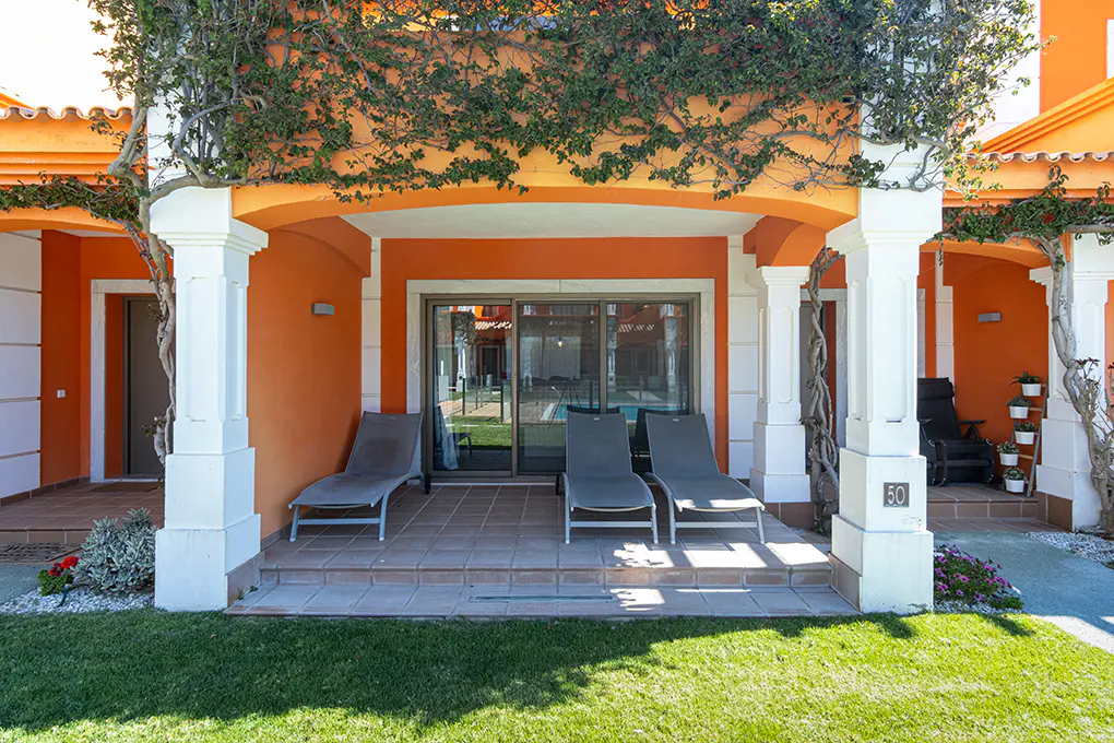 Orange building with white pillars and green vines. Patio with lounge chairs and sliding glass doors. Lawn in foreground.