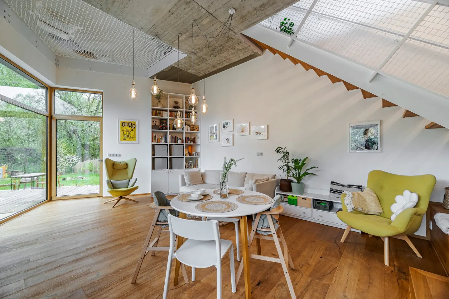 Bright, modern living room with wood floors, white walls, and exposed concrete ceiling. A round table sits near a large window and a green armchair.