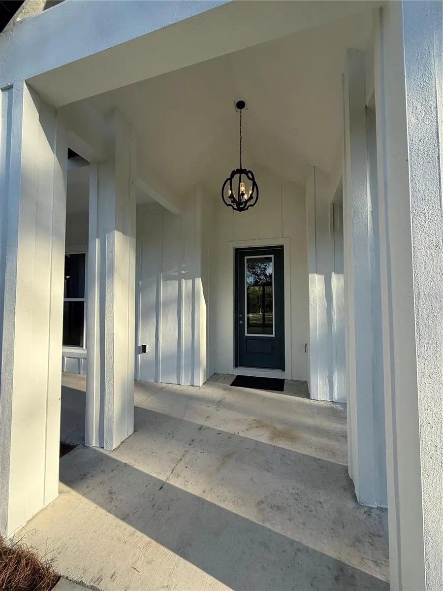 Covered white porch with a dark front door, a black lantern-style chandelier, and concrete flooring.