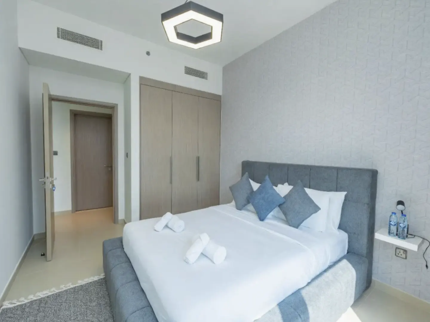 A modern bedroom with a gray bed, white linens, blue pillows, and a geometric light fixture. A closet and open doorway are visible.