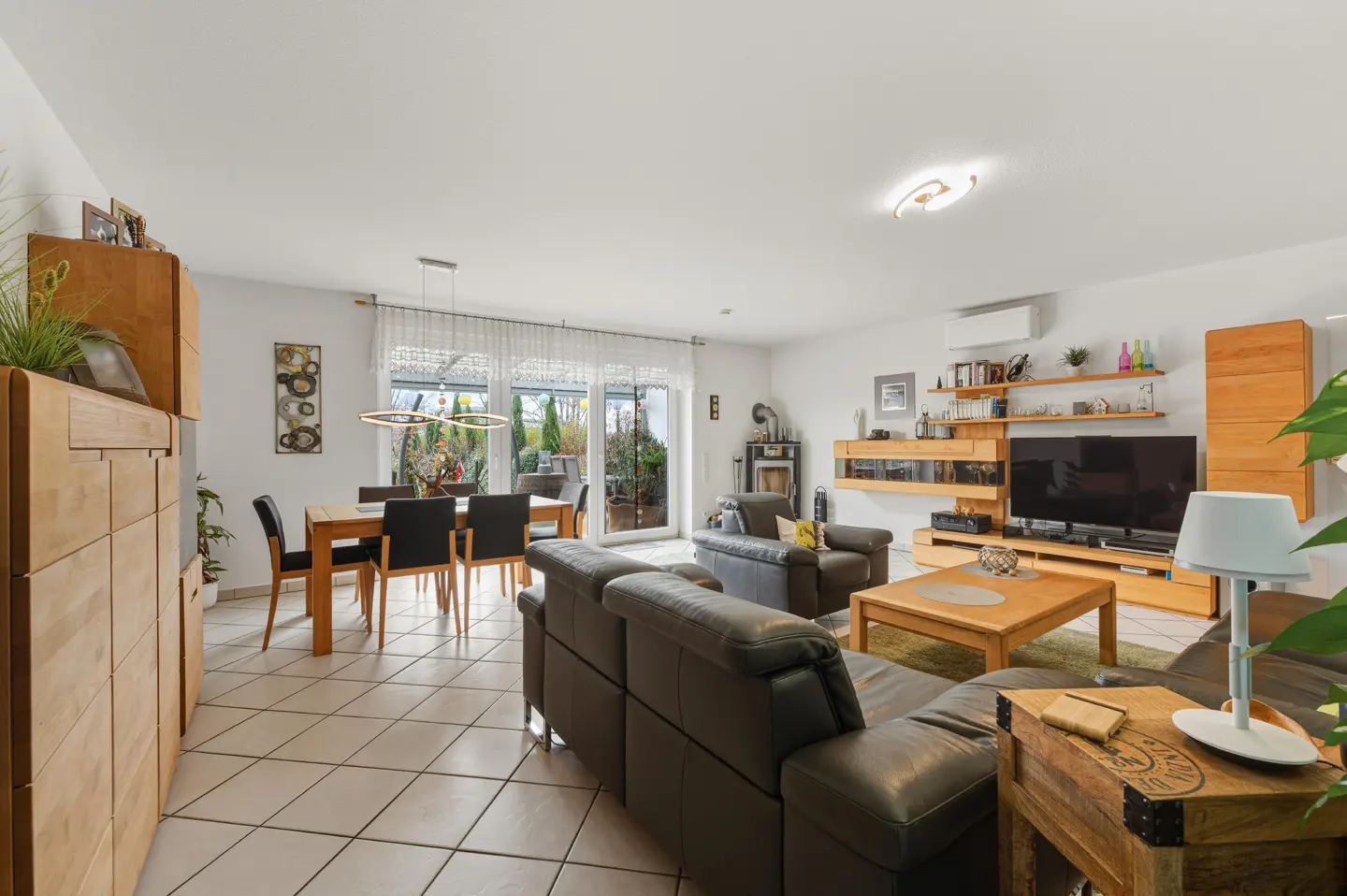 Bright living room with tile floors, a dining table, and a brown leather sofa. A TV and wooden shelves are on the wall.