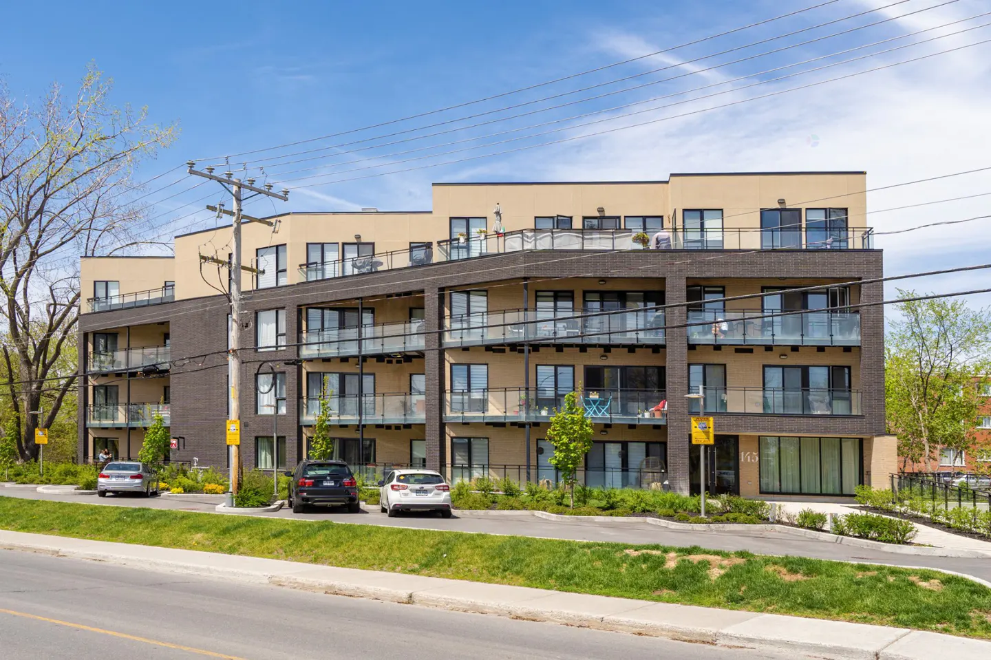 Modern four-story apartment building with tan and dark brick facade, glass balconies, and parked cars in front.