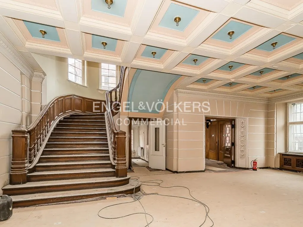 Interior view of a large room with a wooden staircase, coffered ceiling with blue accents, and the Engel & Völkers logo.