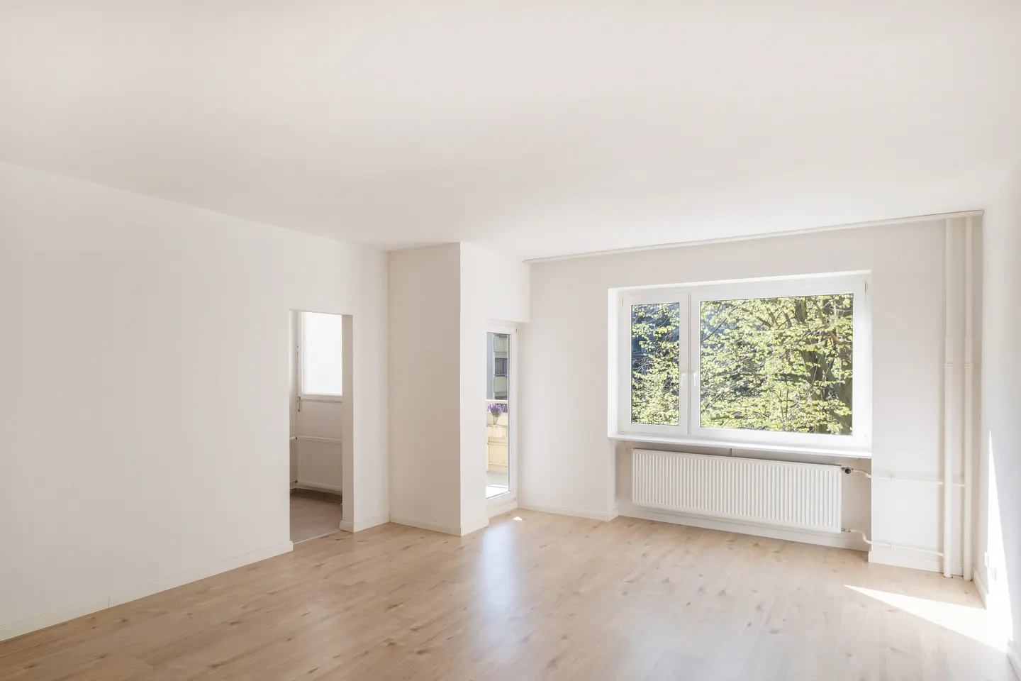 Bright, empty room with white walls, light wood floors, a window with green trees outside, and a doorway to another room.
