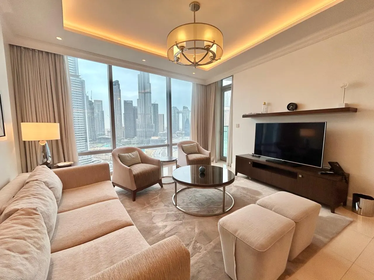 Living room with beige sofa, chairs, and ottomans. Large windows offer a city view. A TV and dark wood console sit against the wall.
