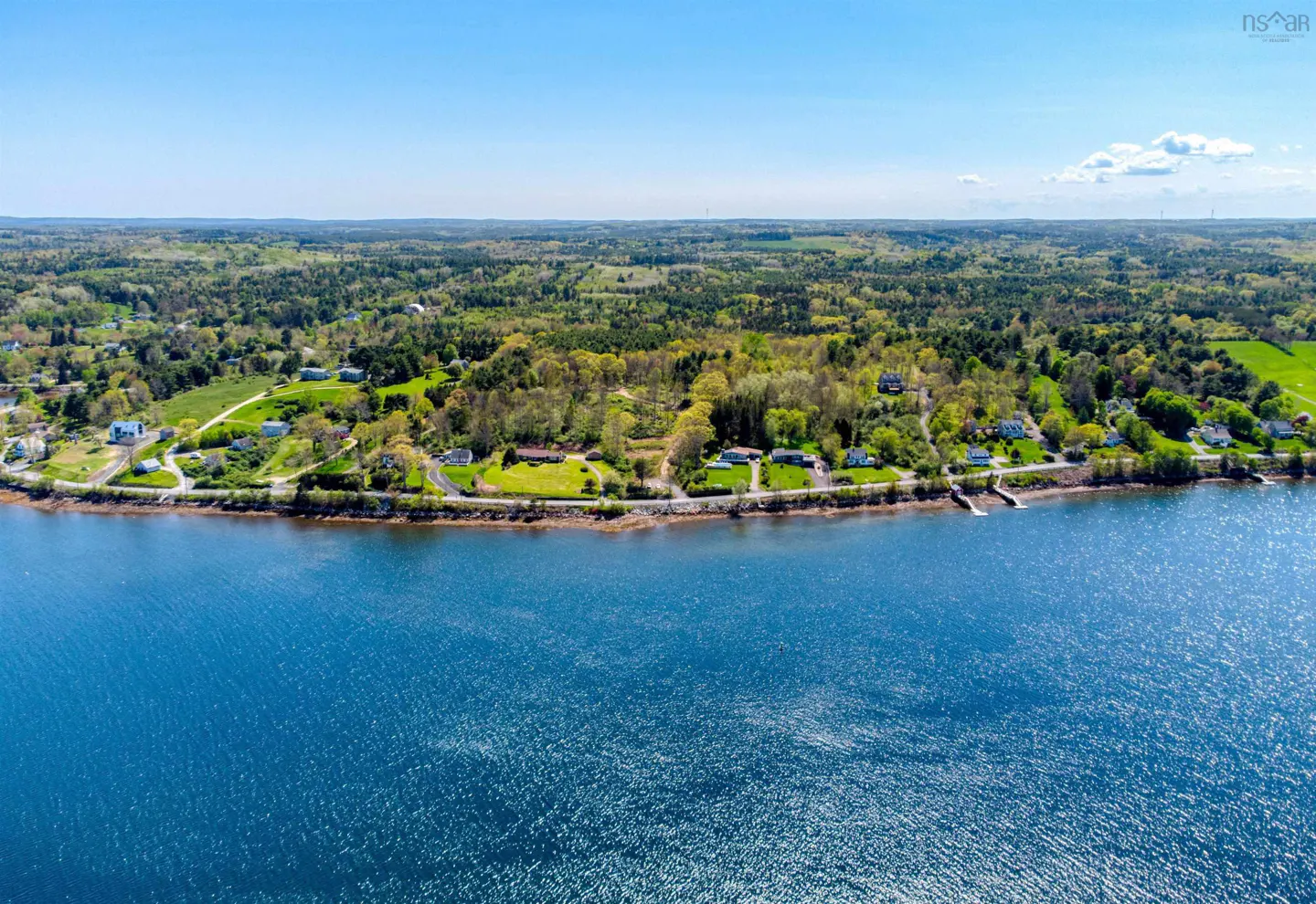 Aerial view of a coastline with blue water, green trees, and houses under a clear blue sky.
