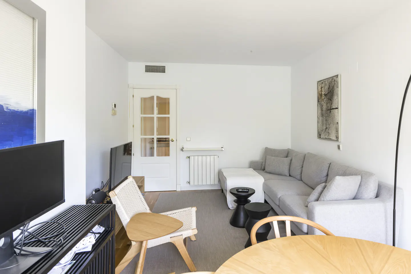 Bright living room with a gray sectional sofa, wood table and chair, black side tables, and a TV. White walls and door.