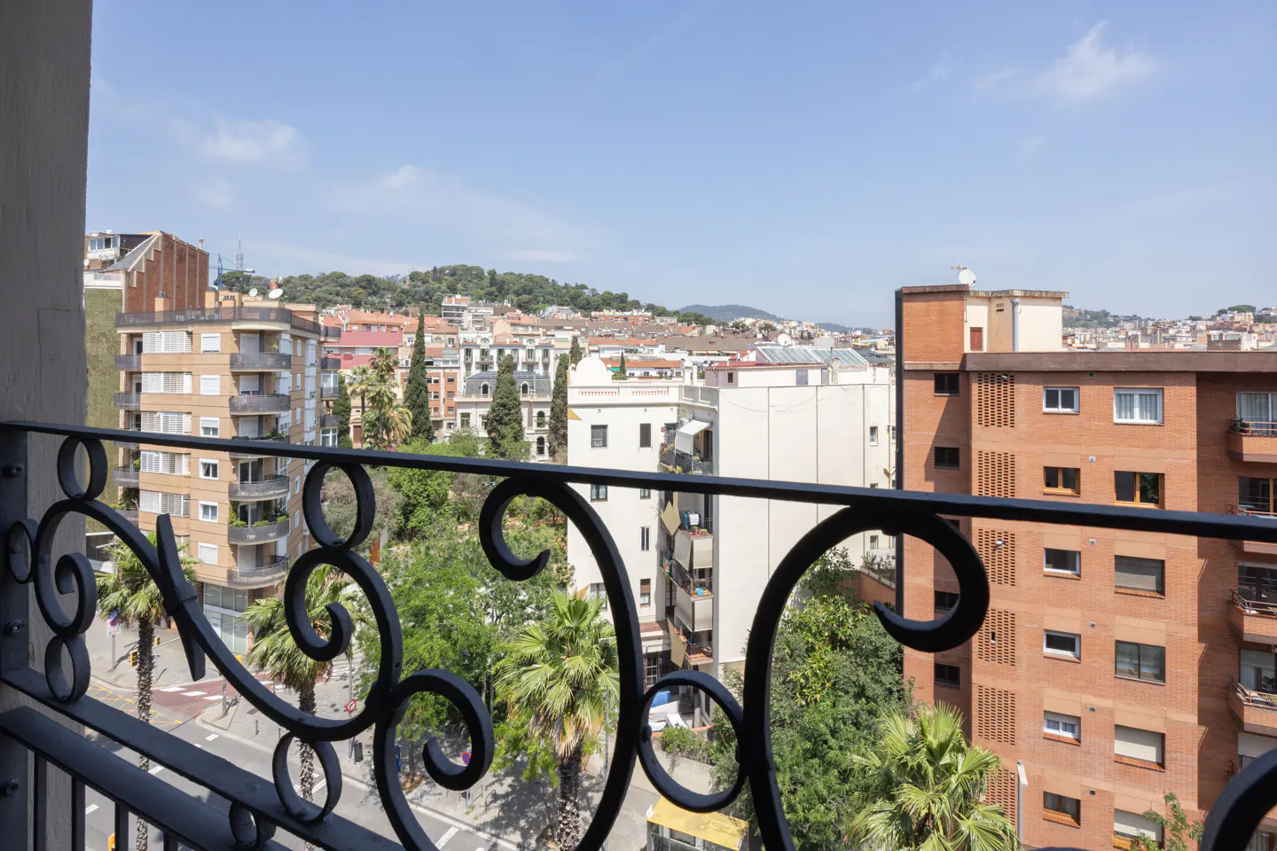 View from a balcony with black wrought iron railing overlooking a city with buildings and trees under a blue sky.