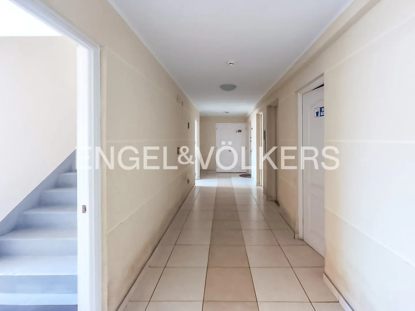A bright, long apartment hallway with beige walls, tiled floors, and white doors, featuring a staircase on the left.