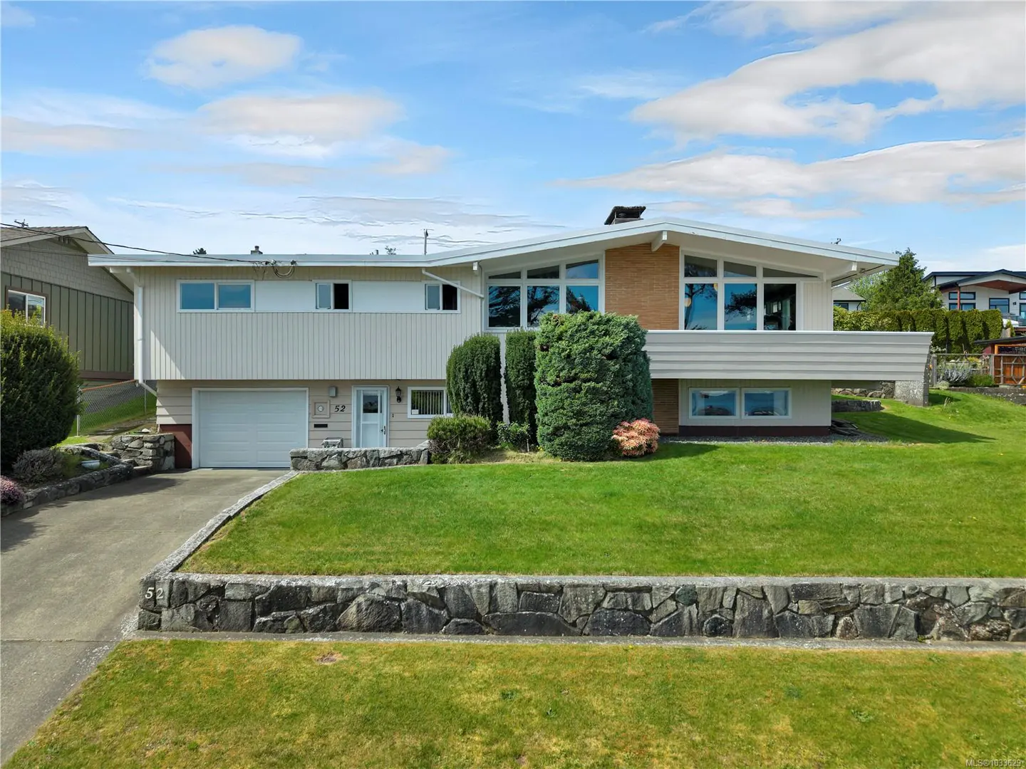 A two-story beige house with a green lawn and stone wall under a blue sky with clouds.