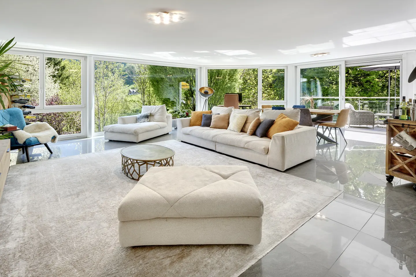 Bright, modern living room with a large beige sofa, matching ottoman, and floor-to-ceiling windows overlooking greenery.