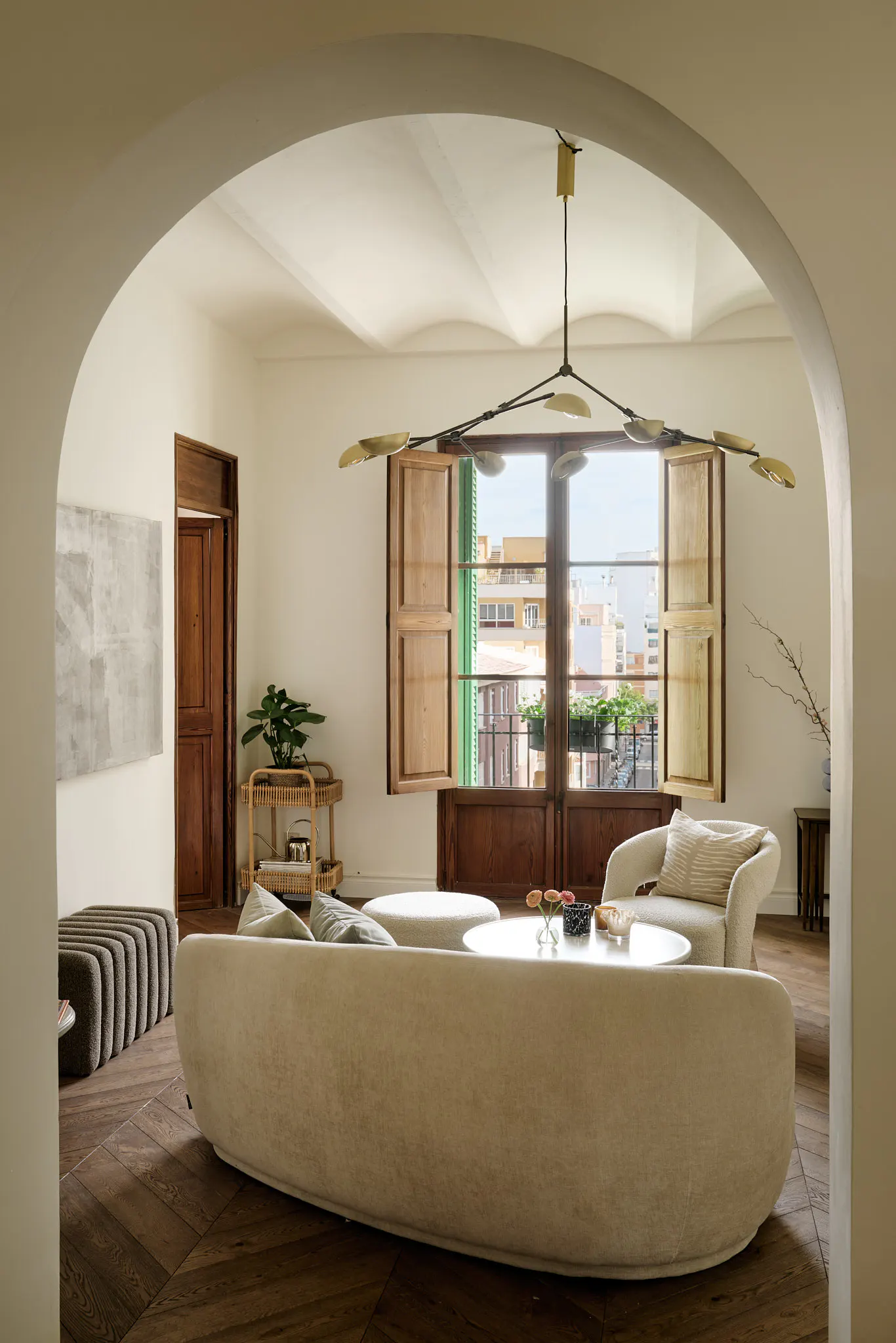 Living room with a beige sofa, round table, and armchair, viewed through an archway. A window with wooden shutters overlooks a cityscape.
