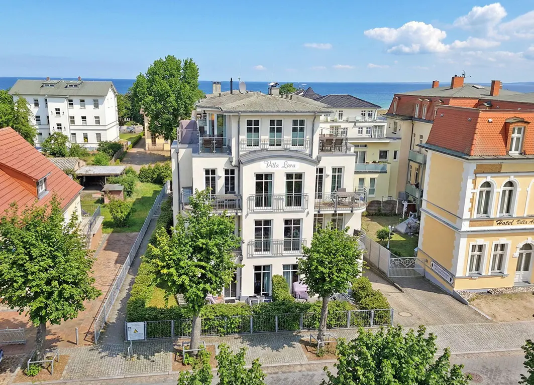 Three-story white Villa Lara building with balconies, surrounded by trees and other buildings, with the ocean in the background.
