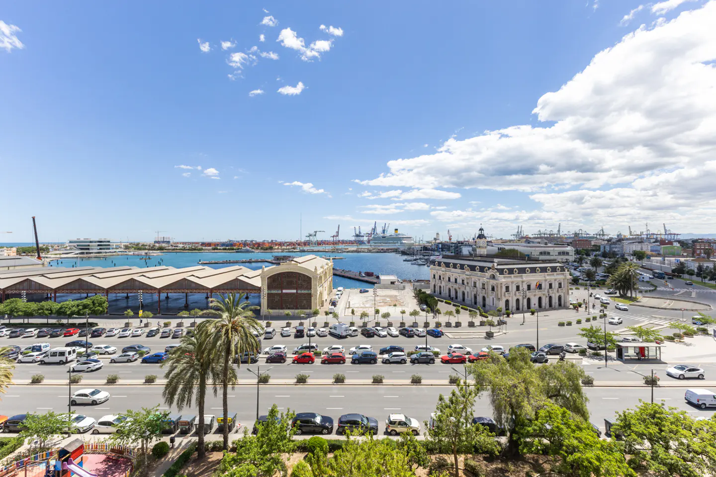 Cityscape view of Valencia, Spain, featuring a harbor with a cruise ship, historic buildings, and a busy street with cars and palm trees.
