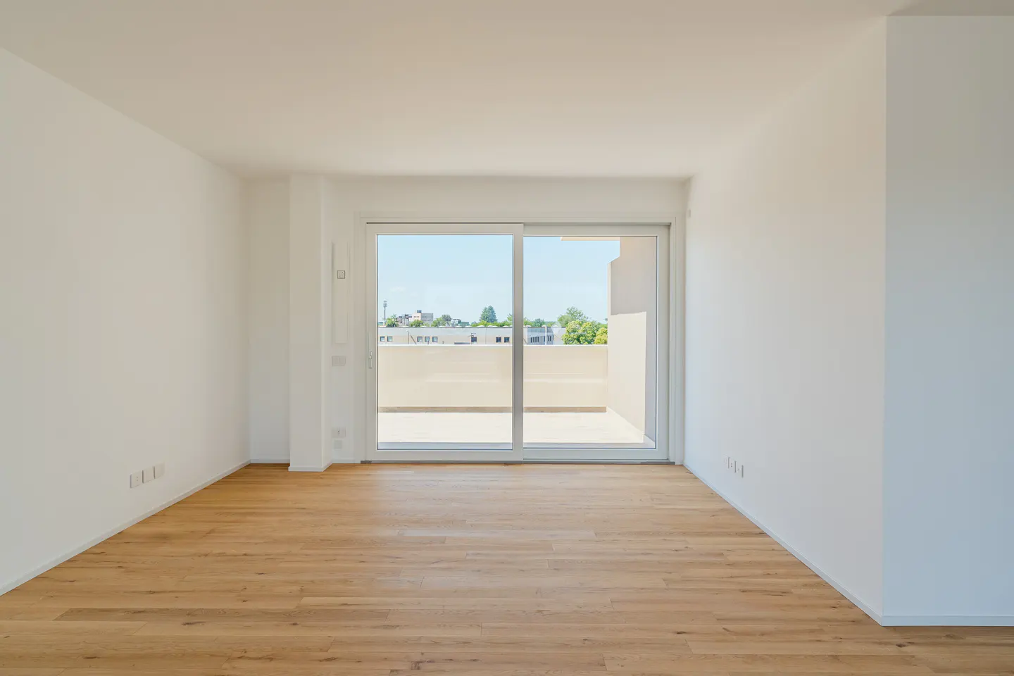 An empty room with white walls and a light wood floor. A sliding glass door leads to a balcony with a view of trees and buildings.