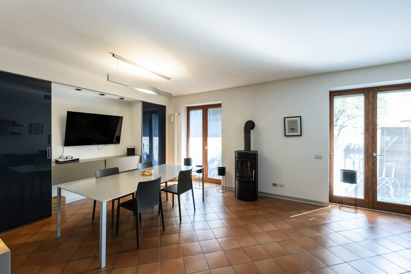 A modern dining room with a white table, black chairs, a TV, and a black wood-burning stove. The floor is tiled in terracotta.