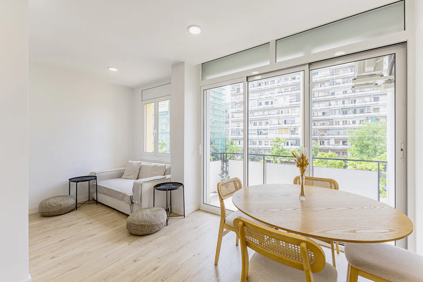 Bright living room with a white sofa, round wooden dining table, and large glass doors opening to a city balcony view.