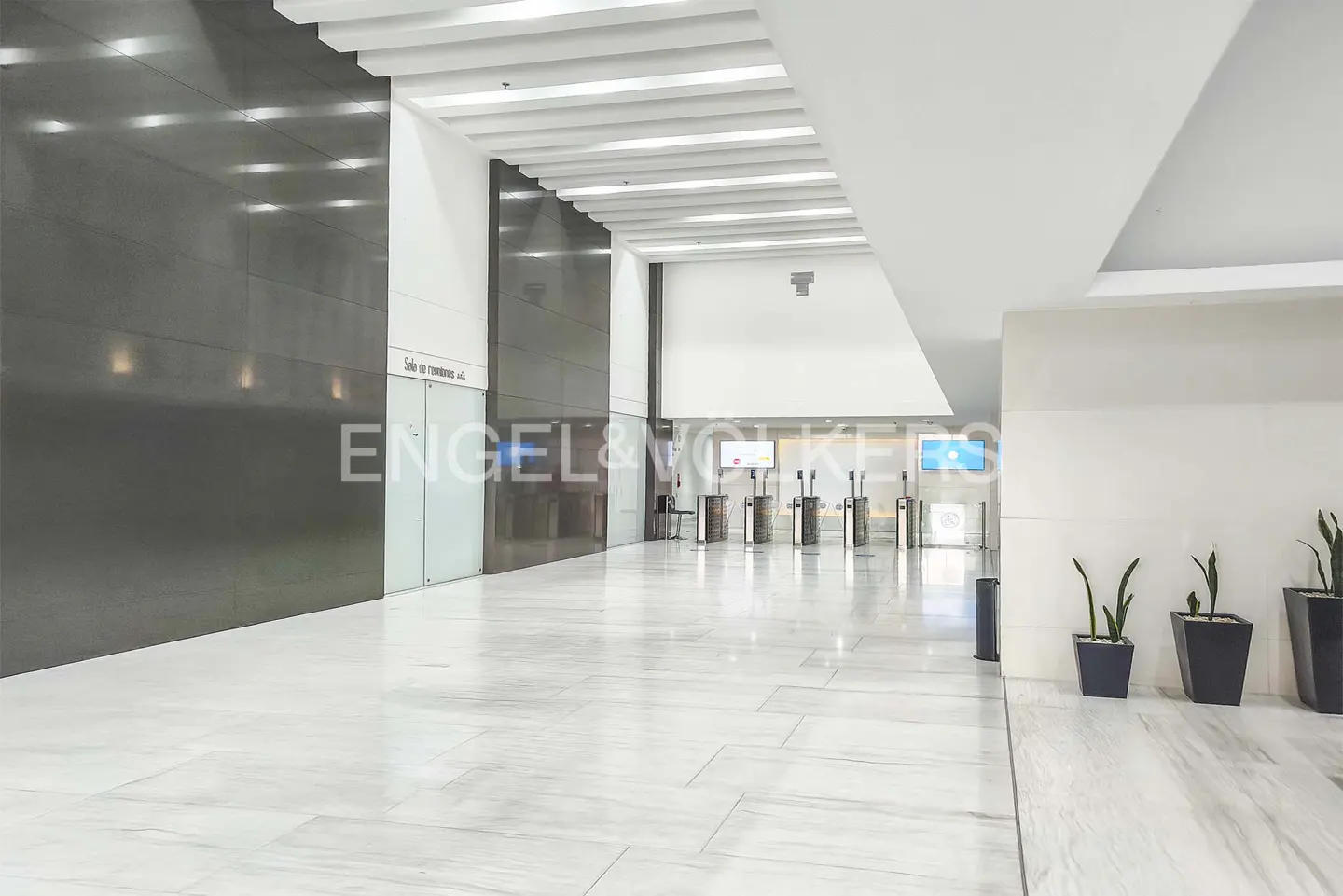 A modern lobby with marble floors, dark metal walls, and white ceilings. Turnstiles lead to a reception area. Potted plants line a wall.