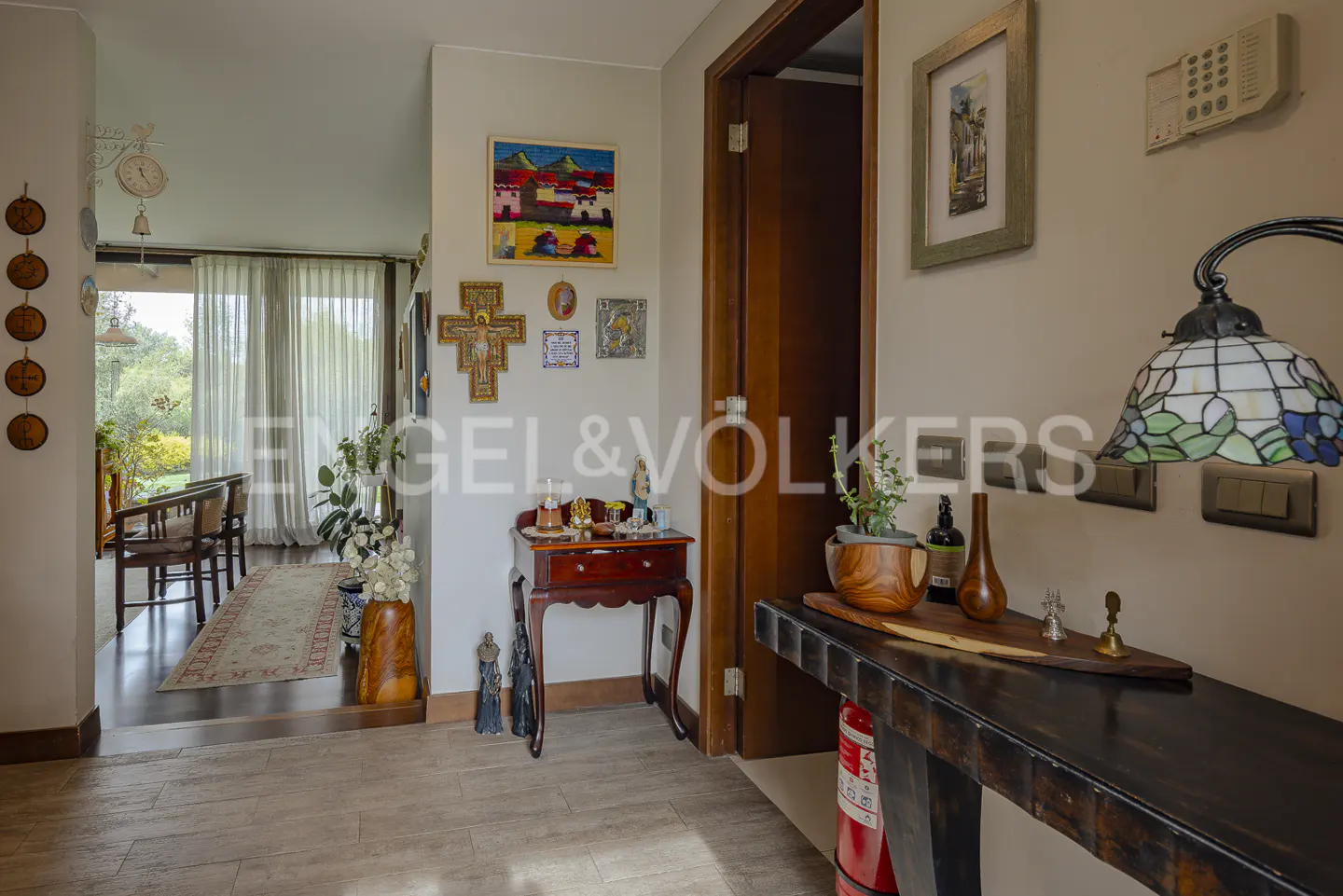 A home's entryway with a dark wood table, a lamp with a blue and green shade, and a view into a living room.