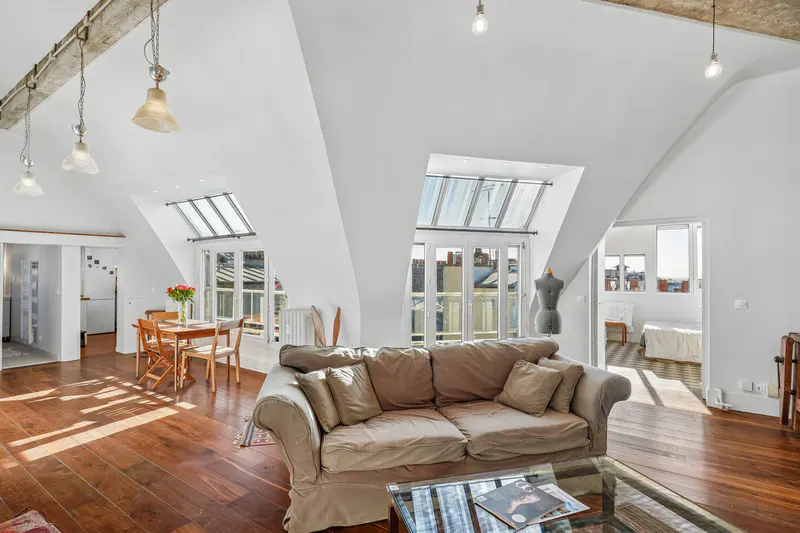 Bright, airy attic apartment with white walls, wood floors, and skylights. A beige sofa sits in the foreground, with a dining table and bedroom visible in the background.