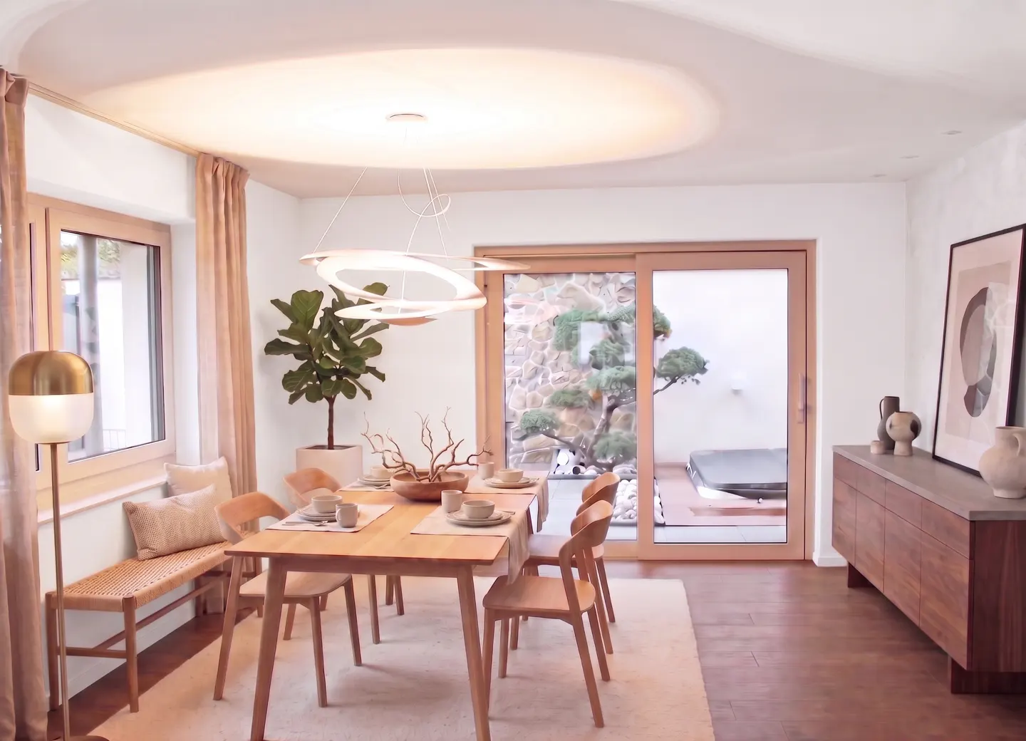 Bright dining room with a wooden table, chairs, and bench. A large window and sliding glass door offer views of a garden. A modern light fixture hangs above.