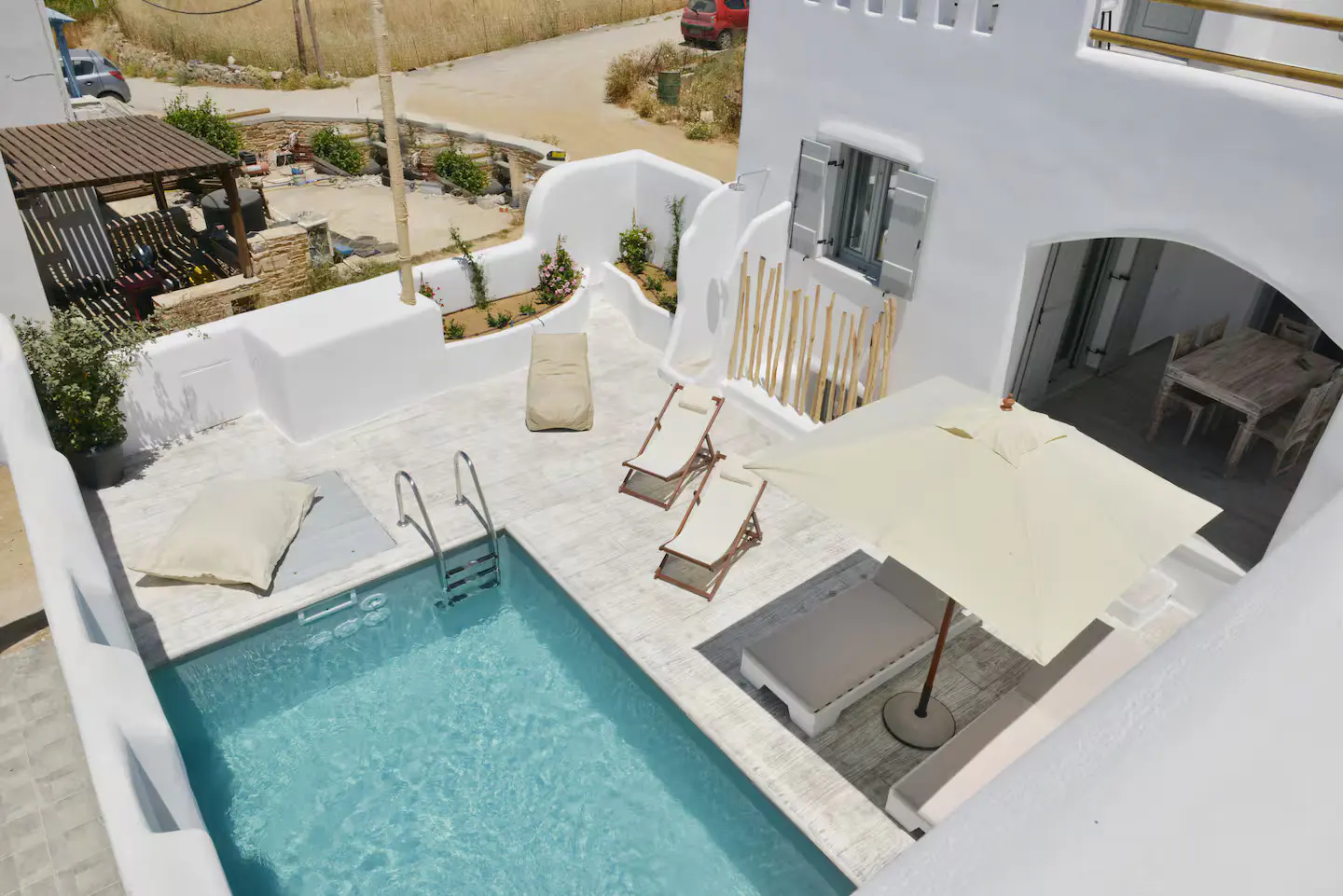 Aerial view of a white villa with a pool, lounge chairs, and a sun umbrella. The pool has clear blue water. A dining table is visible through an archway.