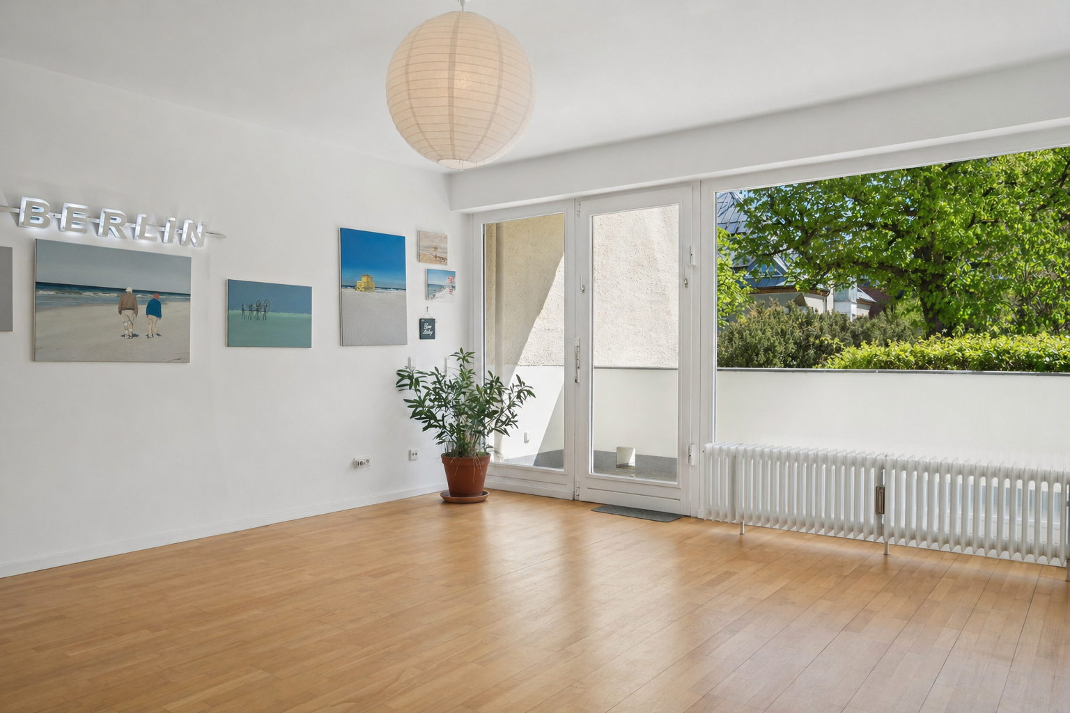 Bright, empty room with wood floors, white walls, and a "BERLIN" neon sign. Paintings hang above a potted plant. A large window shows green trees.