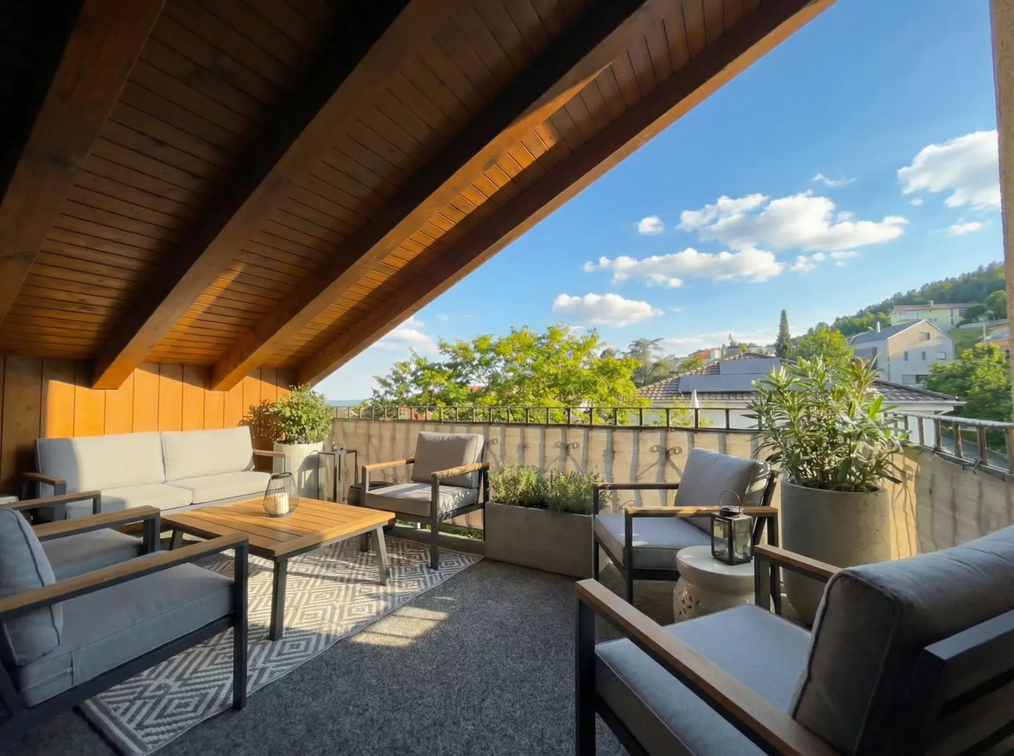 A covered balcony with a wood ceiling, gray furniture, and a view of trees and houses.