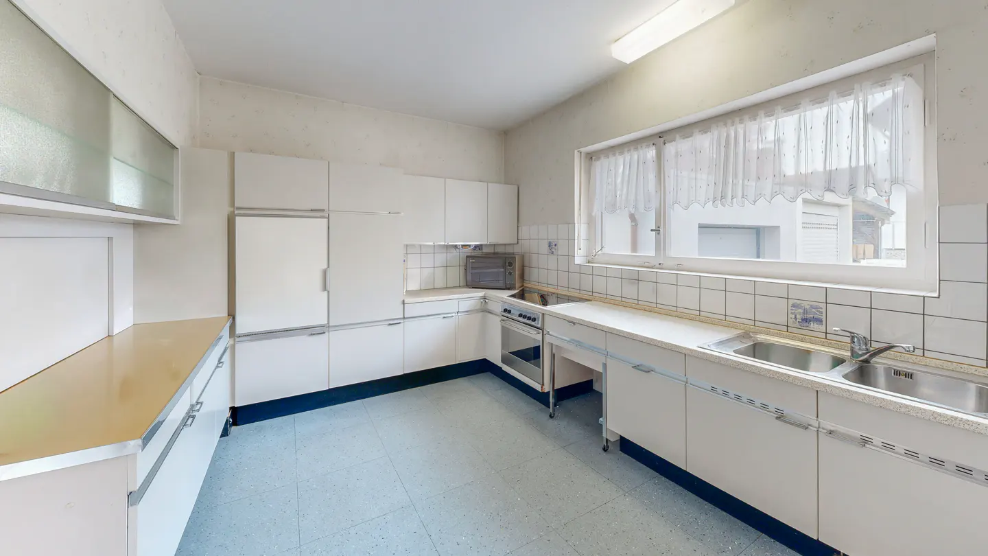 A bright kitchen with white cabinets, a stainless steel sink, and a blue tiled floor. A window with white curtains is above the sink.