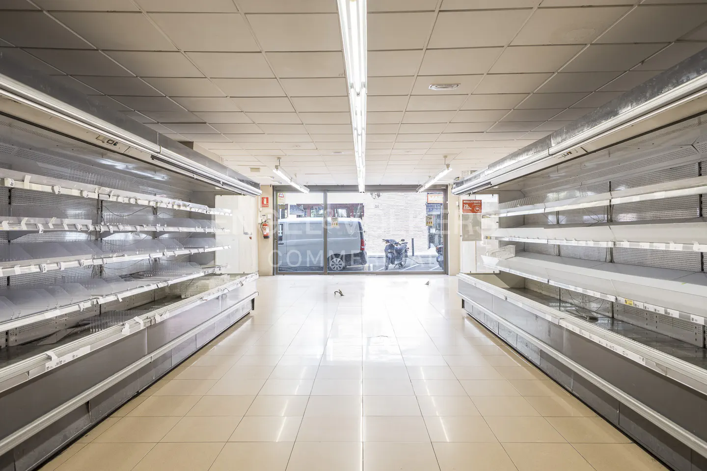 Aisle view of an empty supermarket with bare shelves, a beige tiled floor, and a glass storefront.