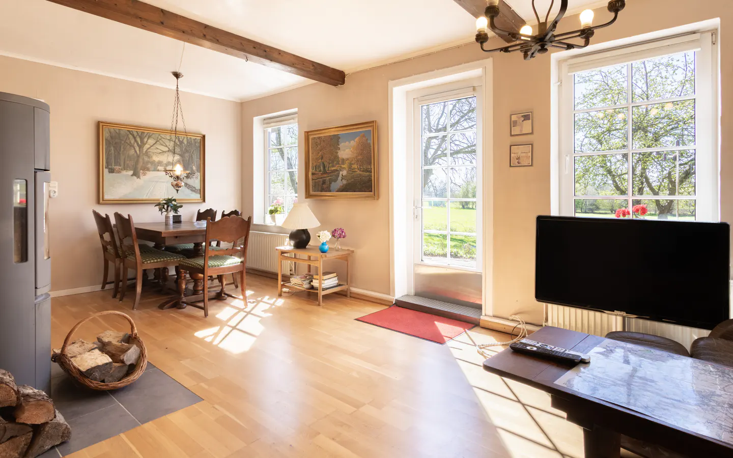 Bright living room with wood floors, dining table, TV, and windows overlooking a green lawn. Sunlight streams in.