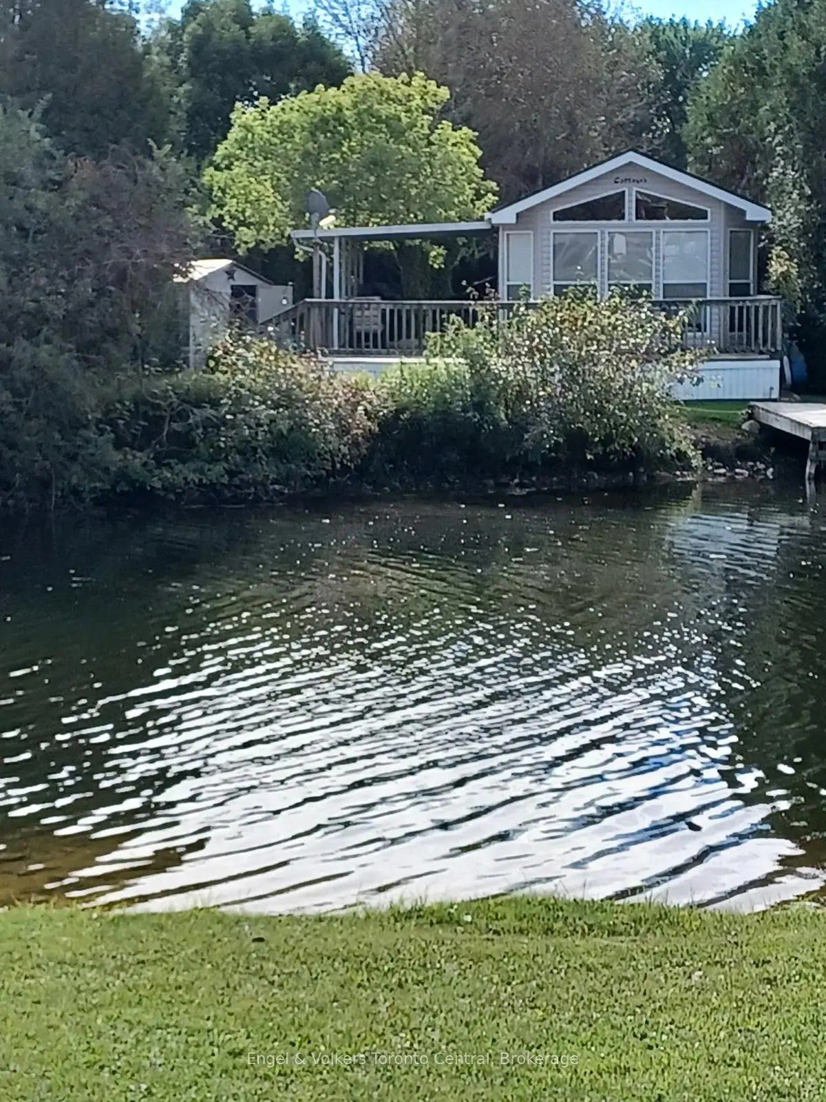 Waterfront property with a light gray house, deck, and dock surrounded by trees and green grass.
